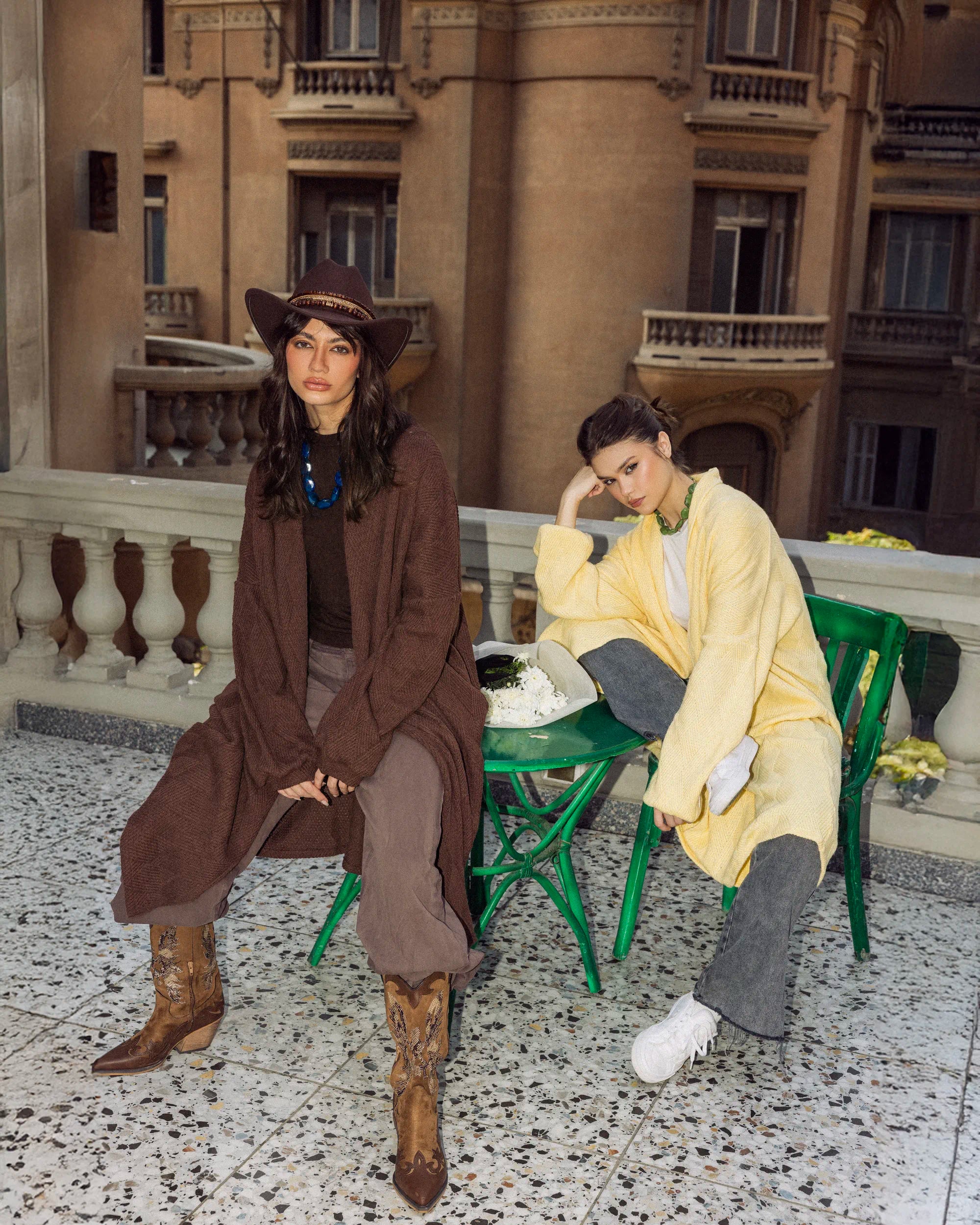 “Two models wearing the Wander Coat in brown and yellow, showing the long relaxed knit silhouette in an outdoor setting.”