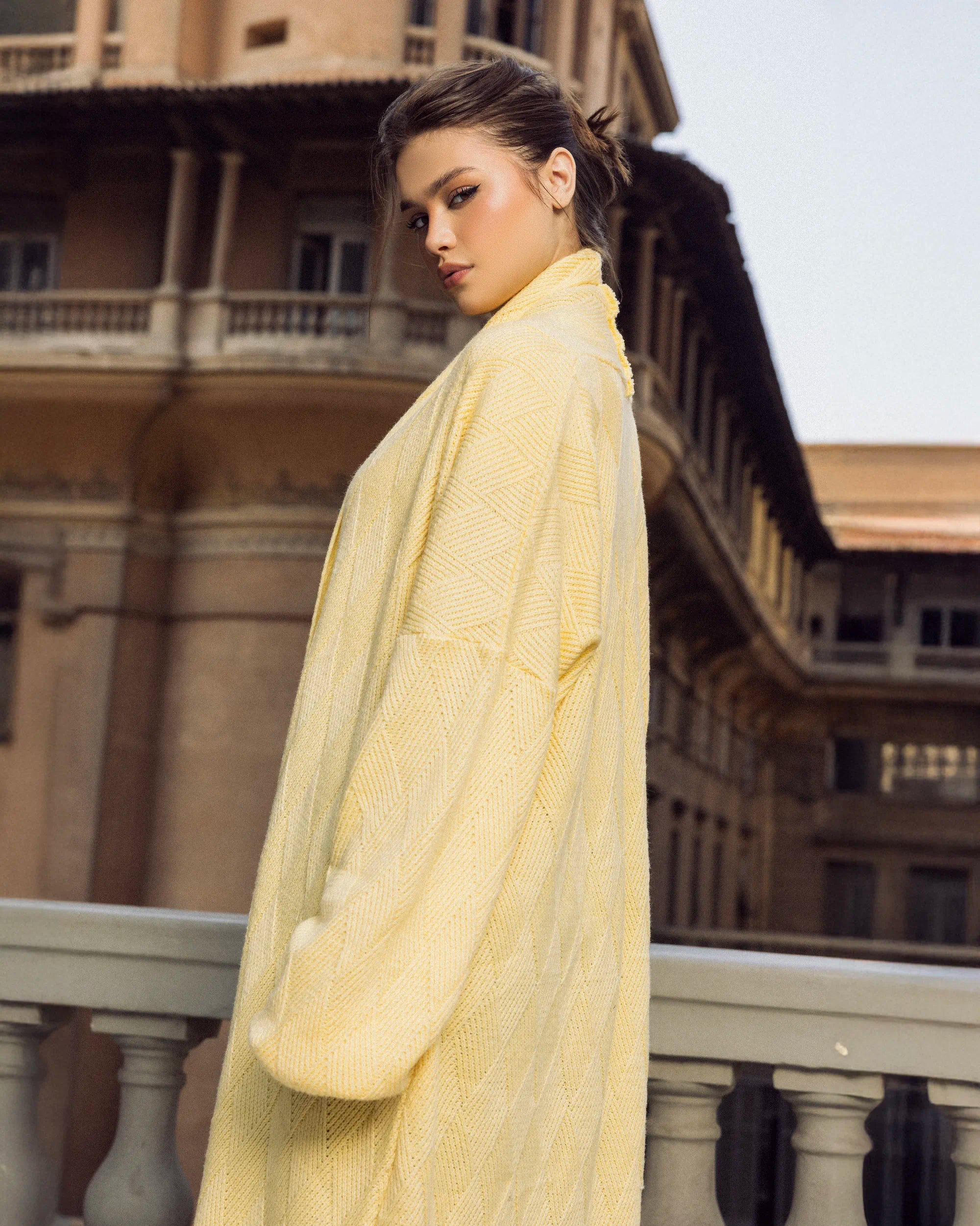 “Side sitting pose showcasing the  Wander Coat’s long draped knit and relaxed winter styling.”