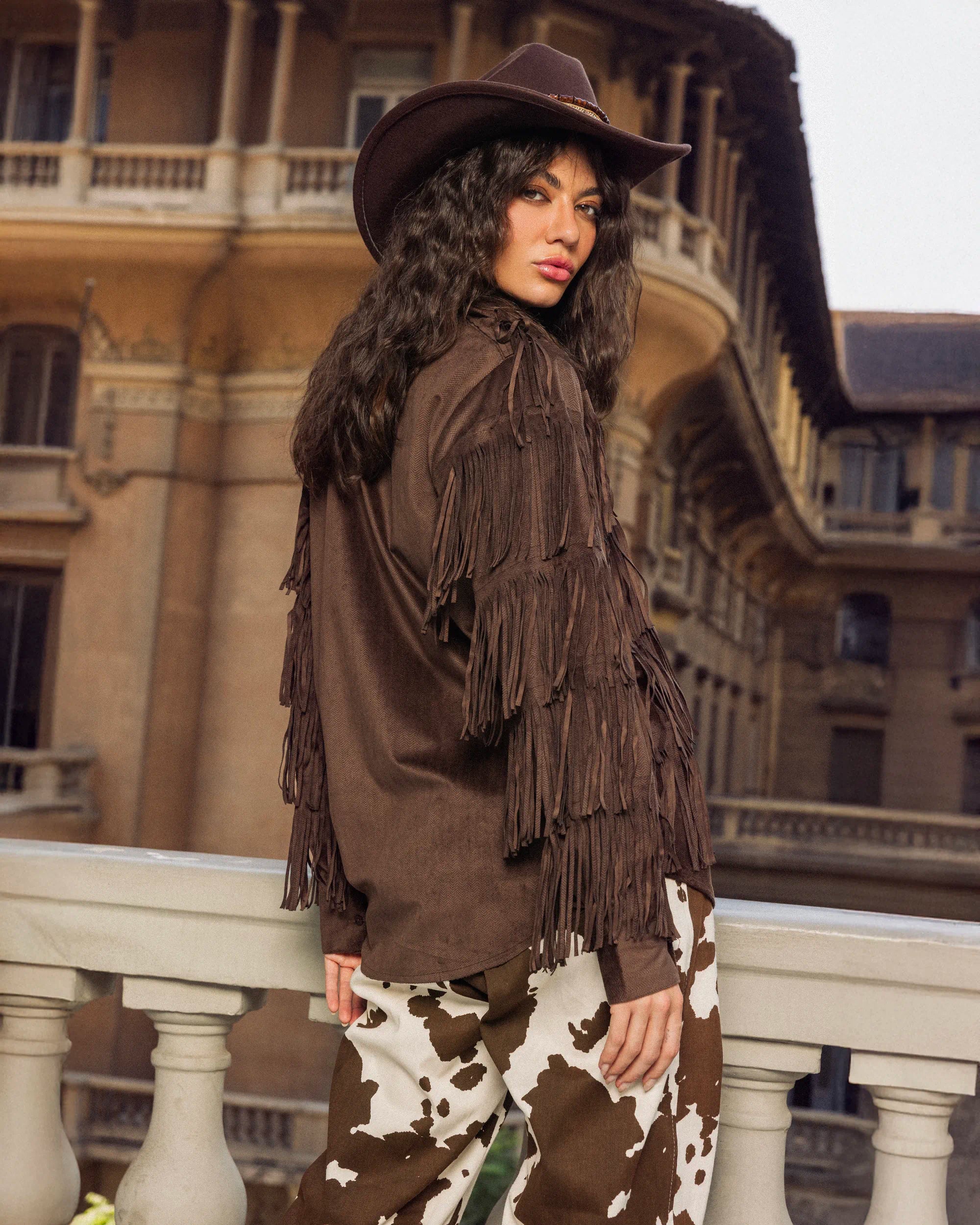 Woman wearing a brown fringed jacket and hat in front of an ornate building.