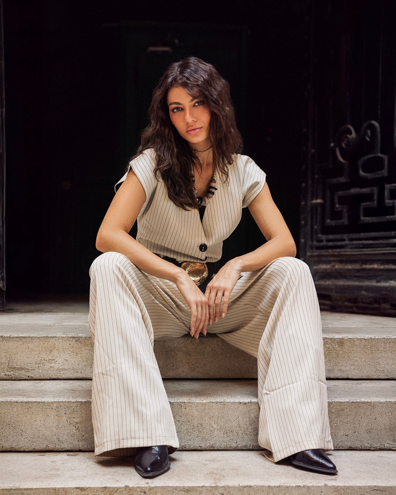 Woman sitting on steps wearing a beige outfit with a belt.