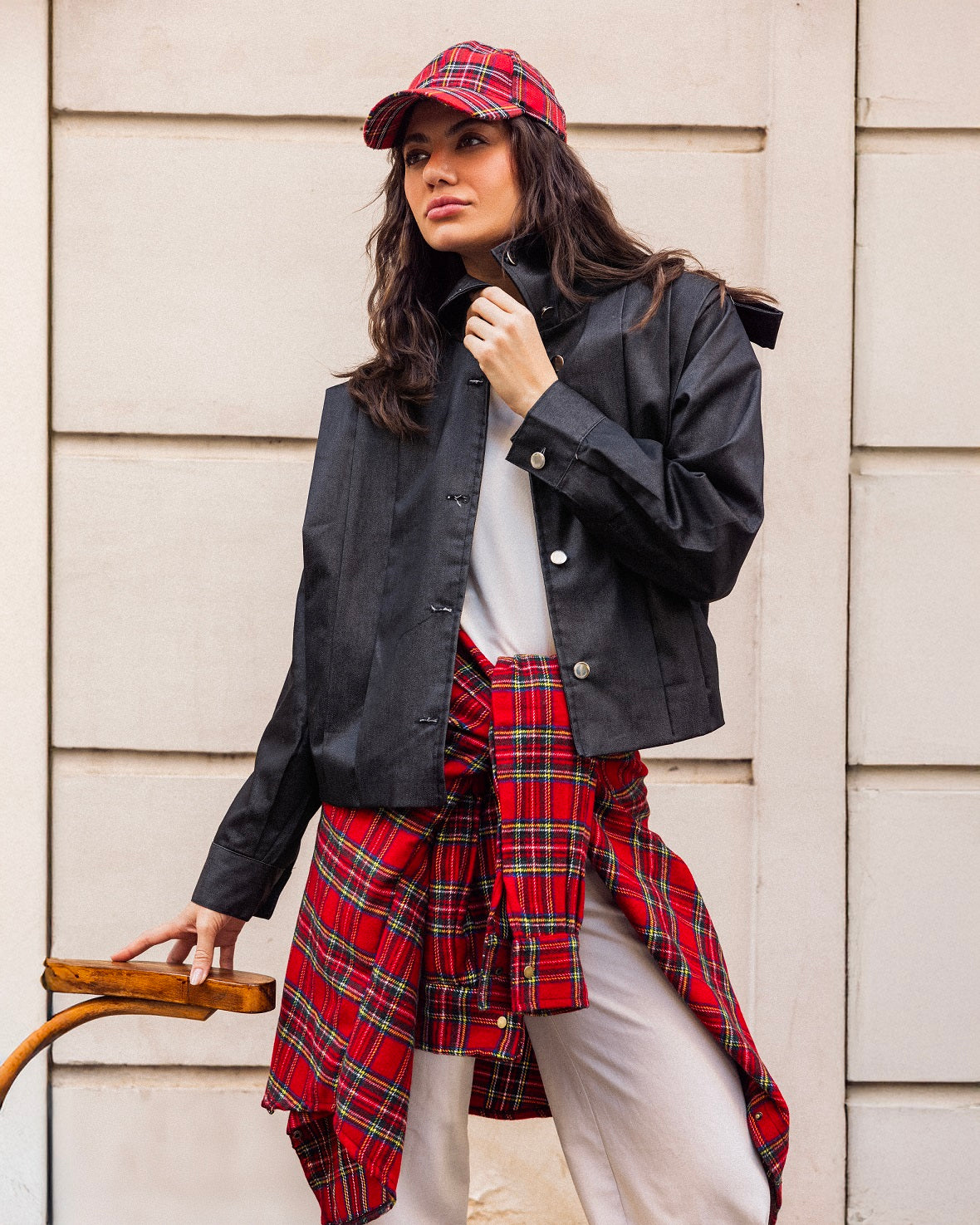 Woman wearing a black jacket, white shirt, and red plaid skirt against a beige wall.