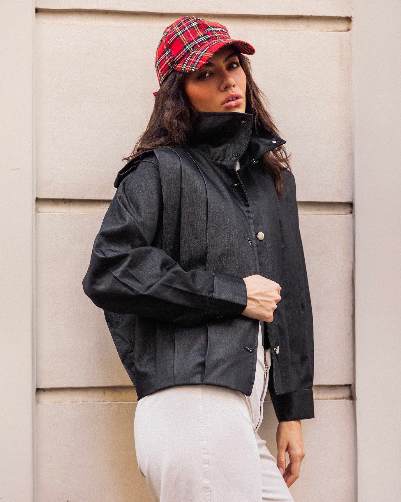 Woman wearing a black jacket and red plaid cap against a beige wall