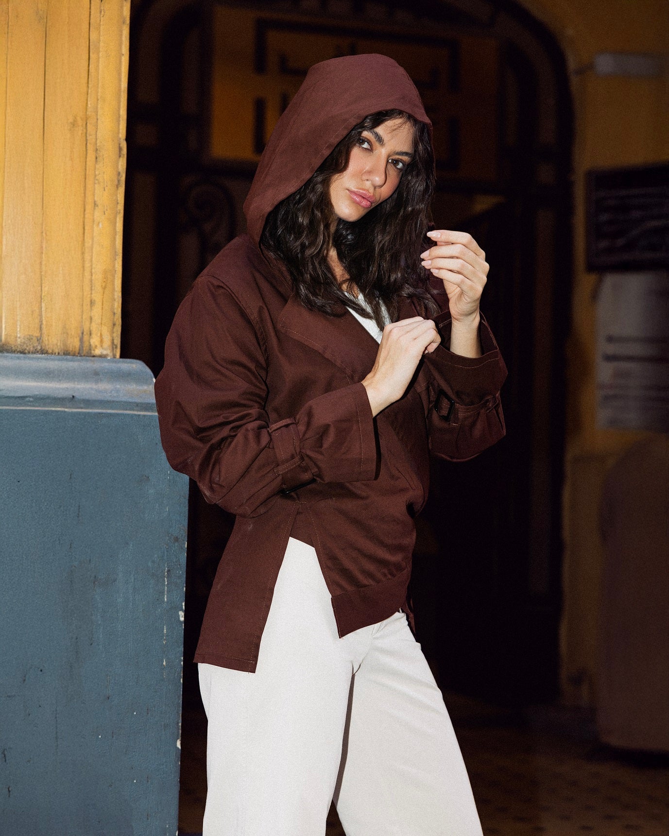 Person wearing a brown hoodie and white pants standing in an industrial setting.