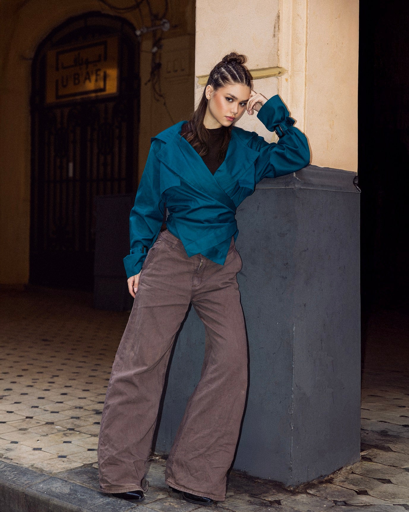 Woman in teal blouse and brown pants leaning against a wall in an urban setting