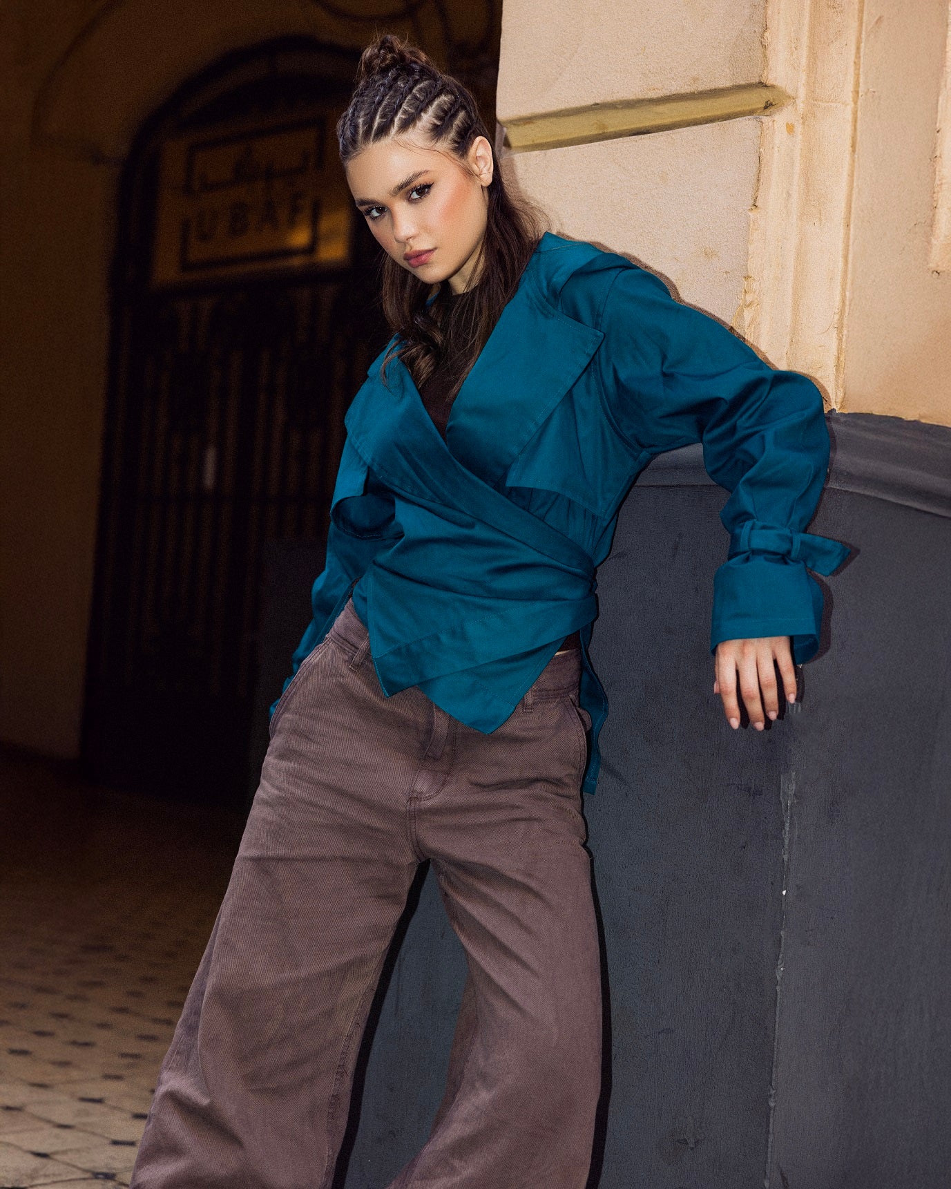 Woman wearing a blue top and brown pants leaning against a wall.