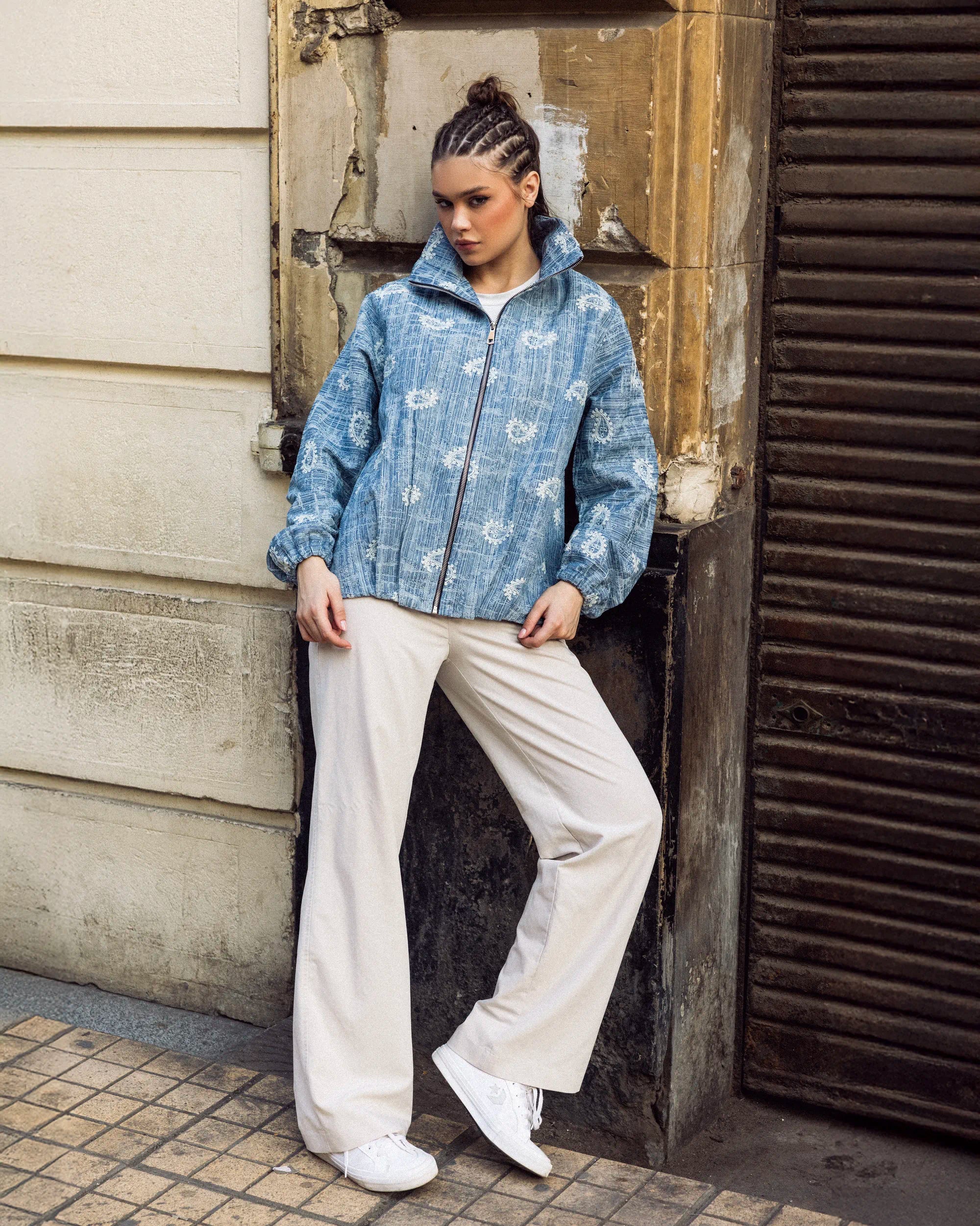 Person wearing a blue floral jacket and white pants standing against a textured wall.
