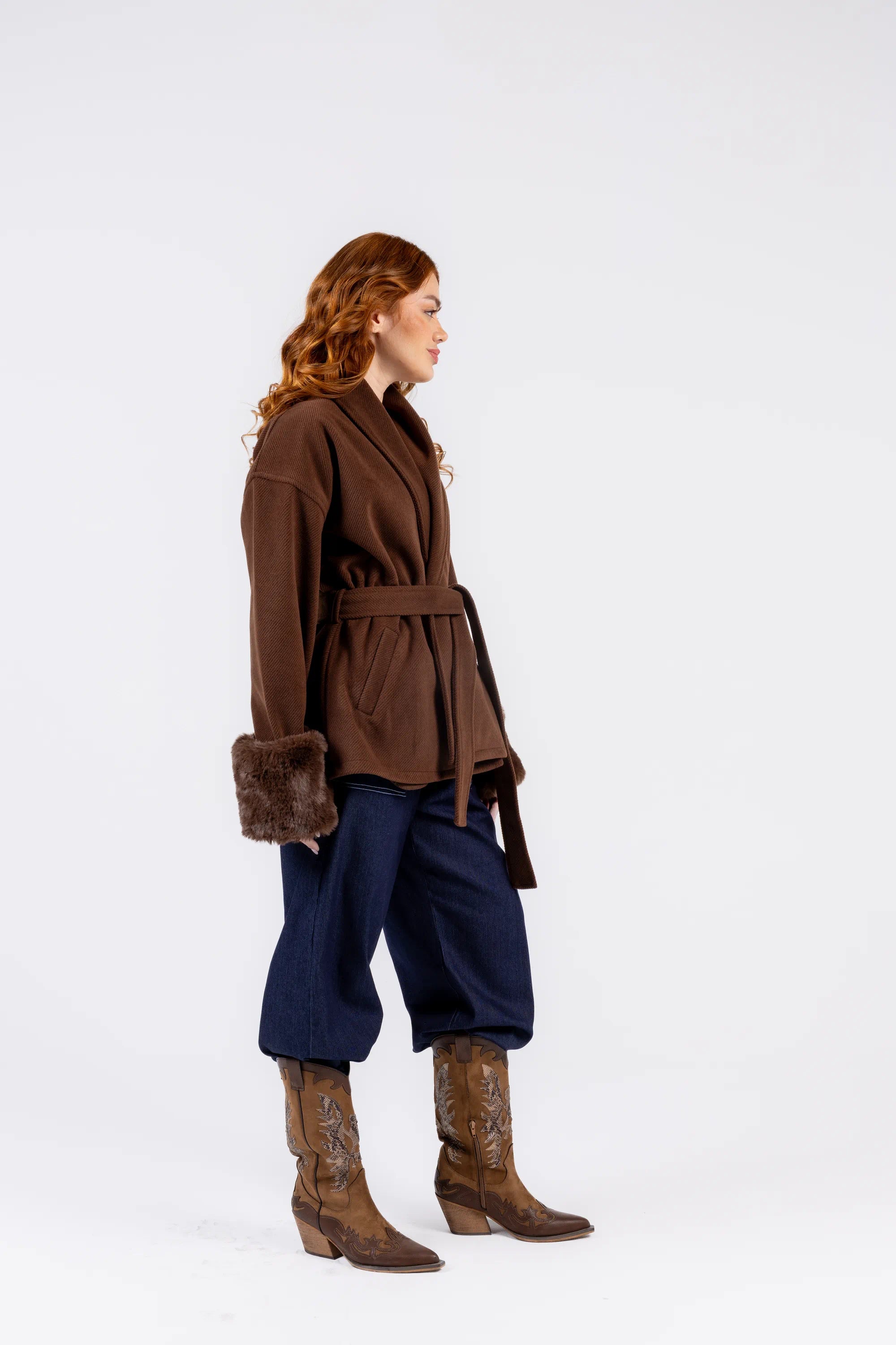 Person wearing a brown coat with fur trim, blue pants, and brown boots on a white background
