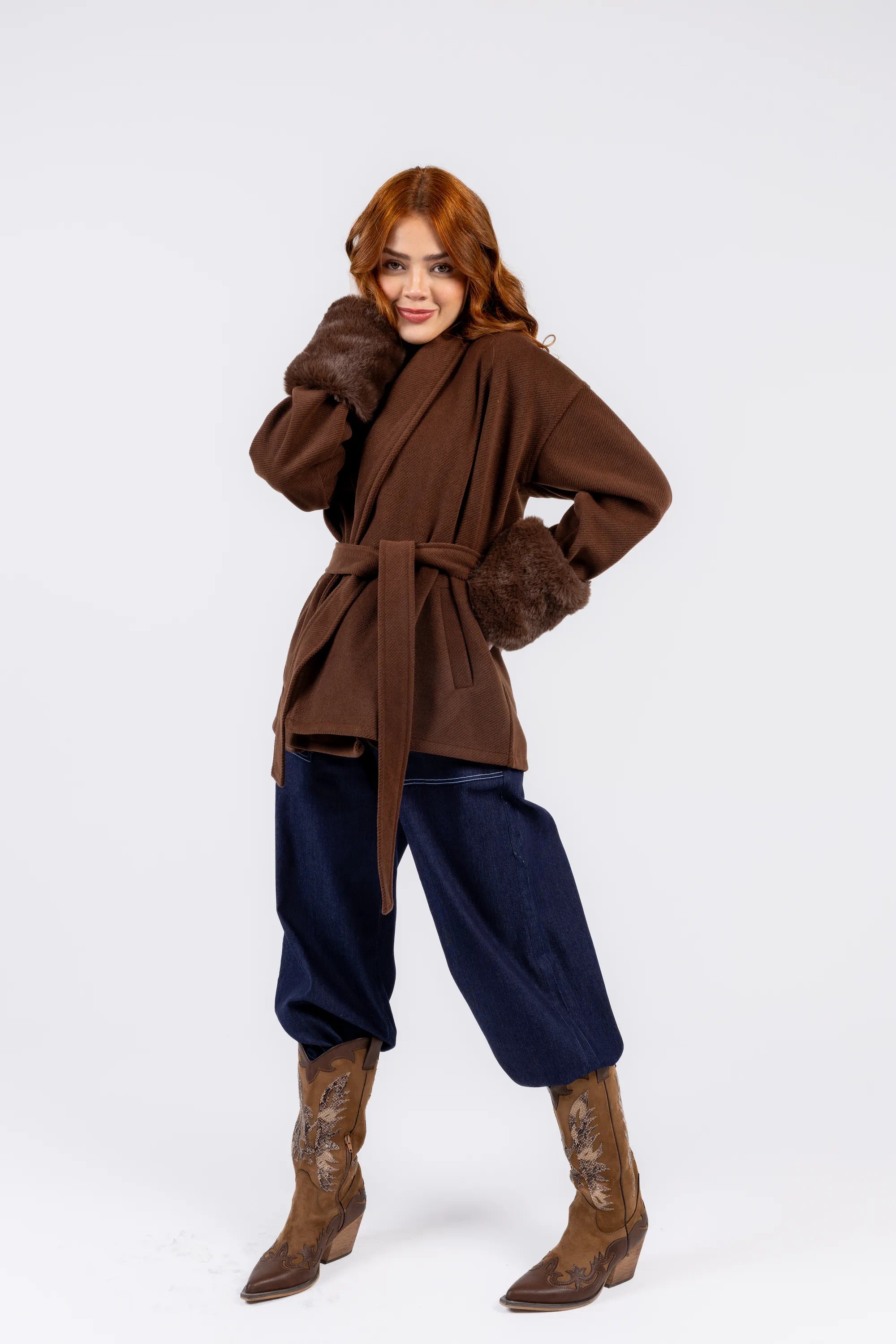 Woman wearing a brown coat with fur trim, navy pants, and brown boots on a white background