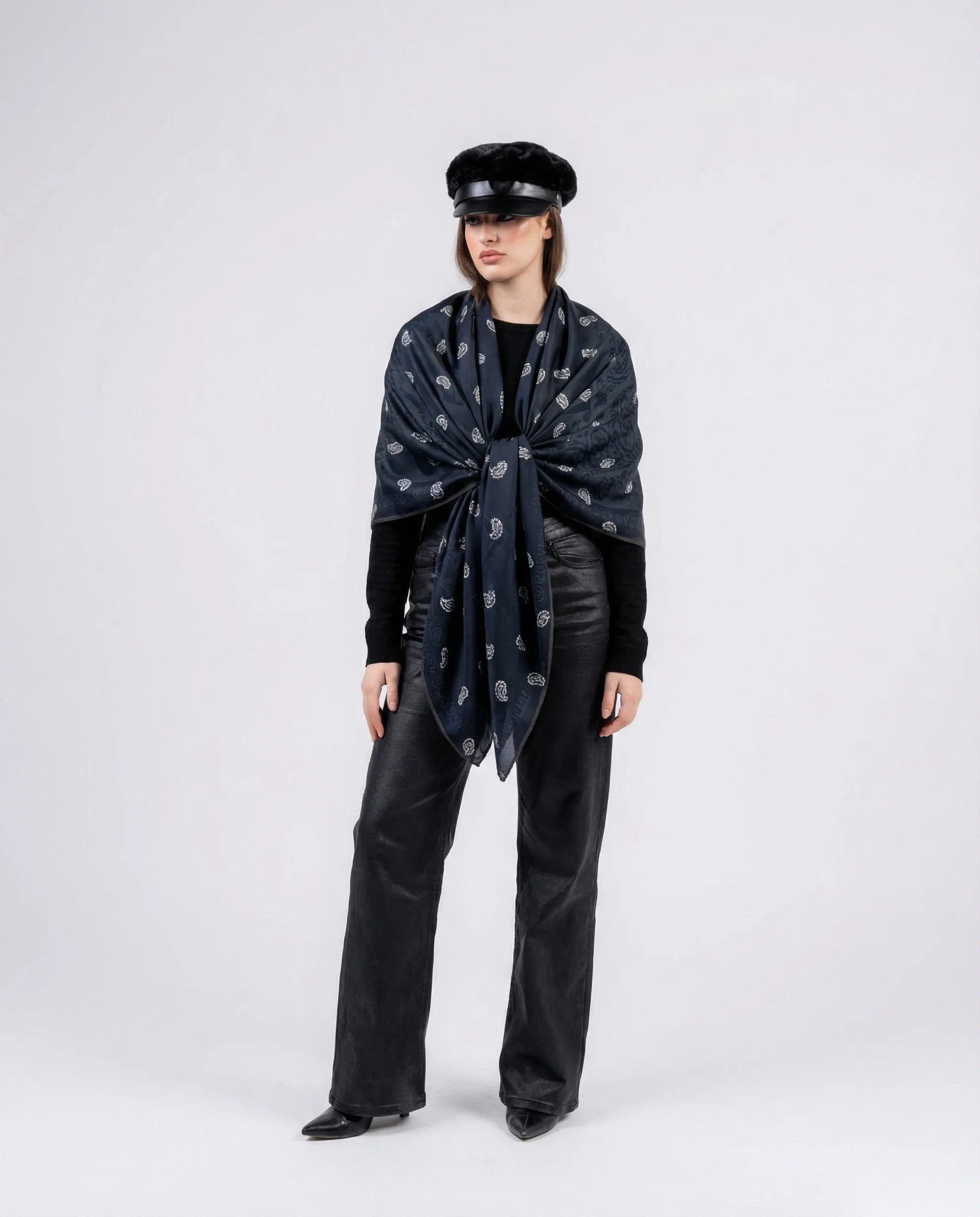 Person wearing a dark scarf with a pattern, black outfit, and hat on a white background