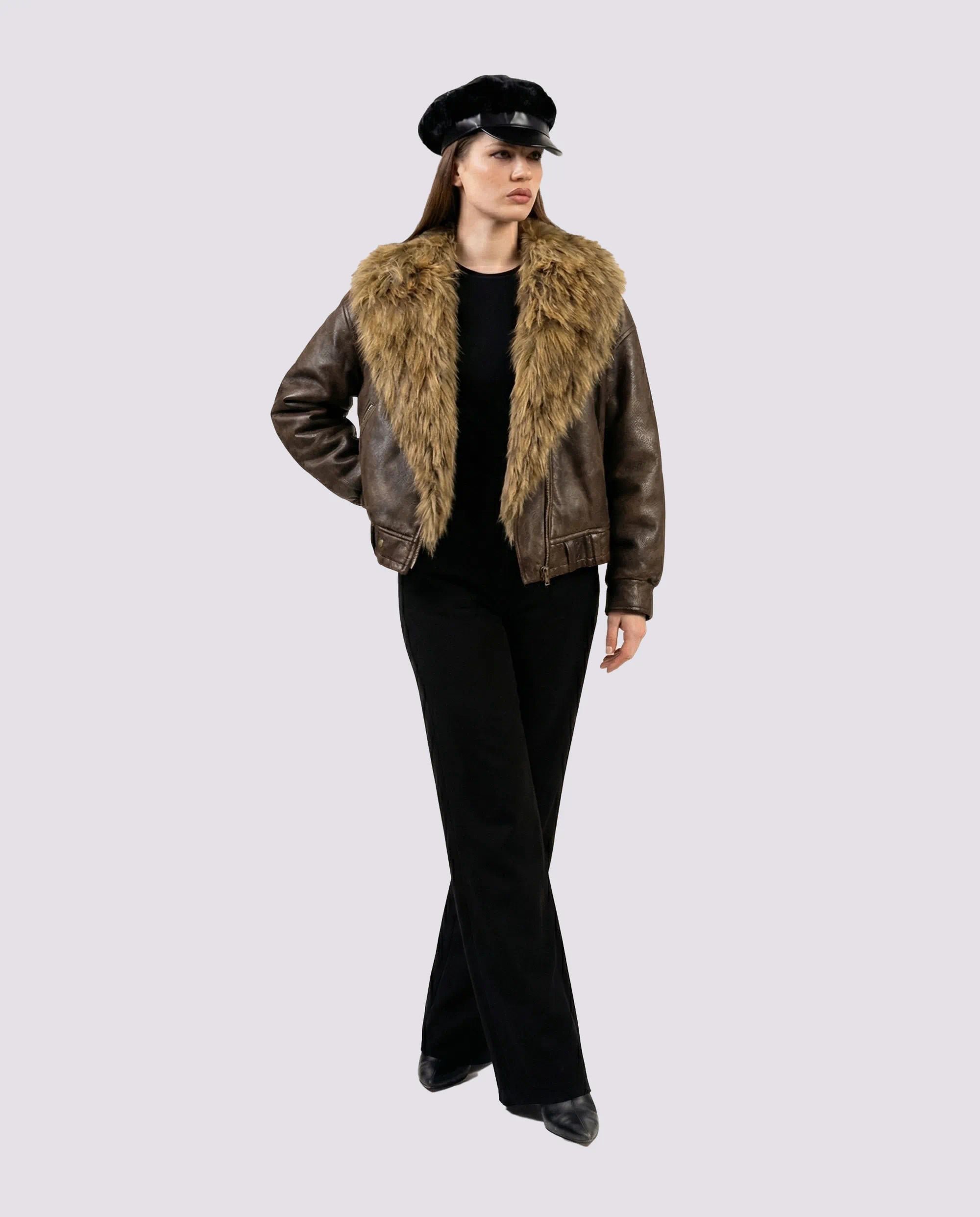 Woman wearing a brown leather jacket with fur collar and black pants on a light gray background